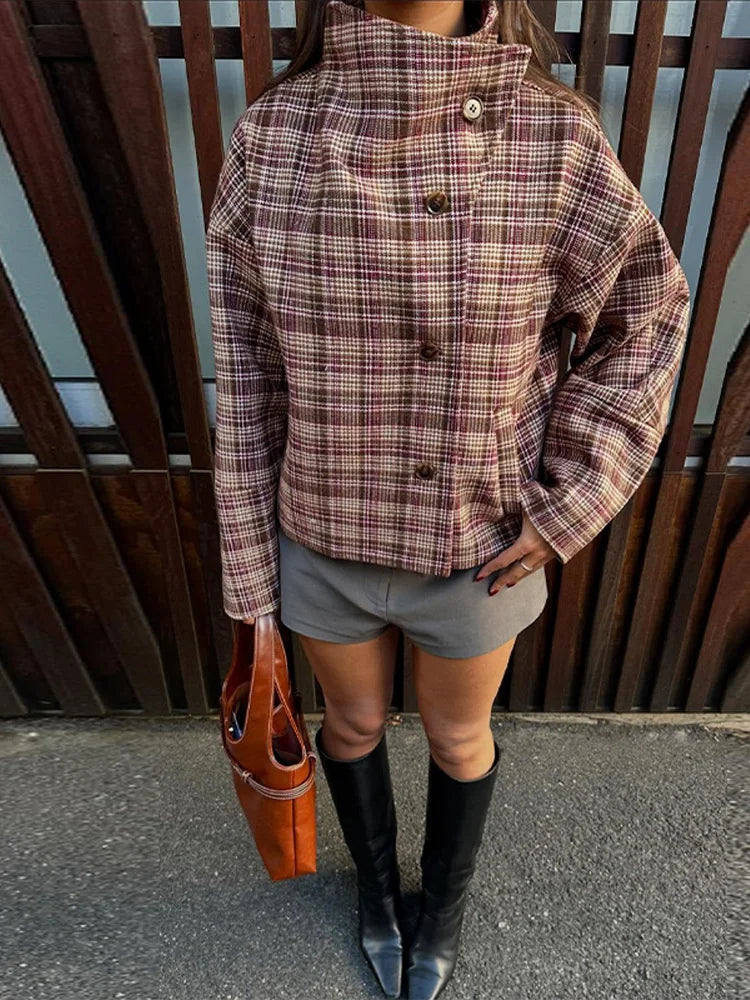Person wearing a plaid jacket, gray shorts, black knee-high boots, and holding an orange bag against a wooden background.