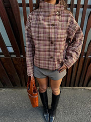 Person wearing a plaid jacket, gray shorts, black knee-high boots, and holding an orange bag against a wooden background.