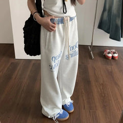 Person wearing white sweatpants with blue text and blue sneakers in a room.