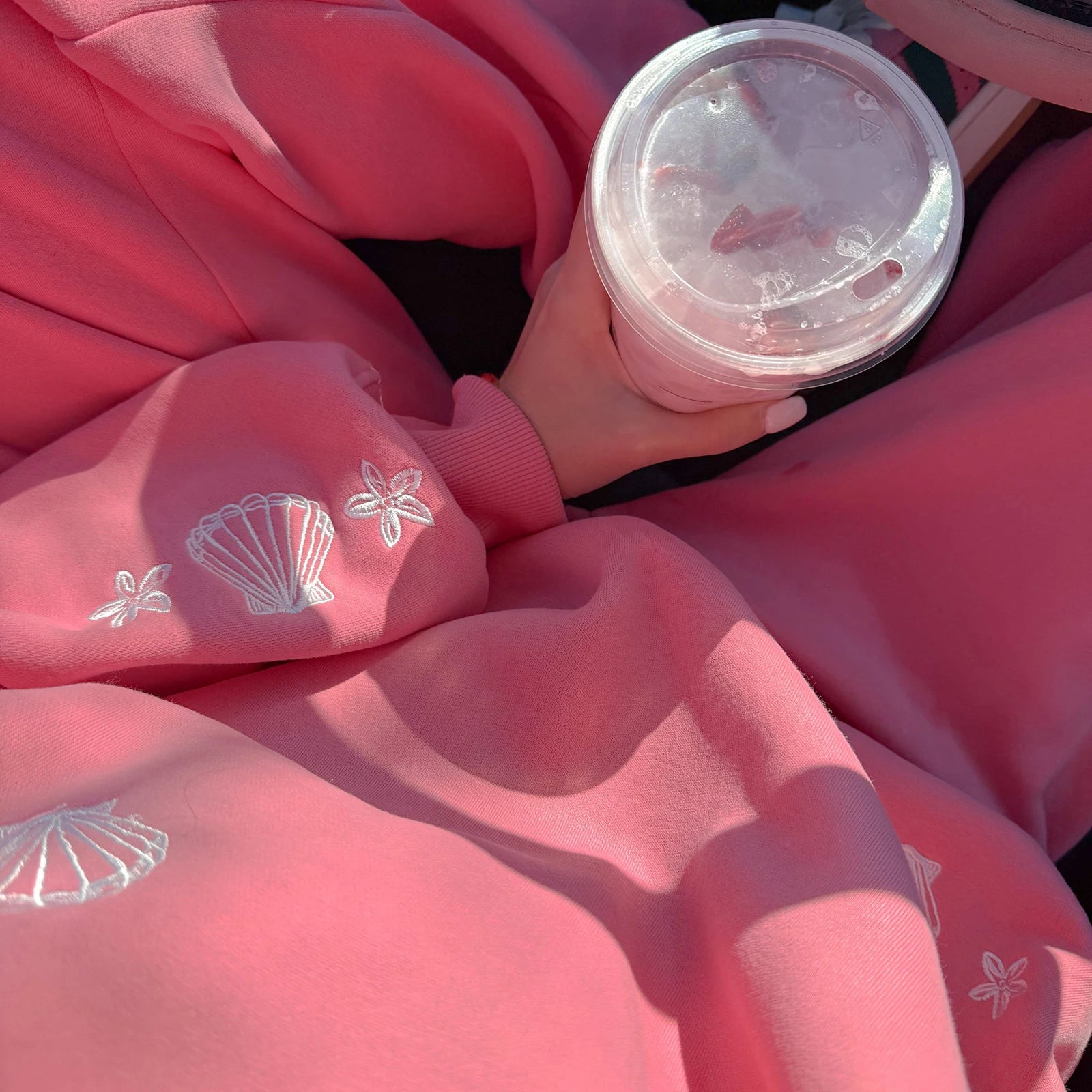 Person wearing a pink sweatshirt with shell designs, holding a clear plastic cup.