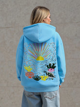 Person wearing a blue hoodie with a colorful ocean-themed design on the back, standing against a plain background.