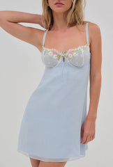 A woman wearing a light blue camisole nightgown with floral lace detailing on the bust, photographed against a plain light gray studio background.