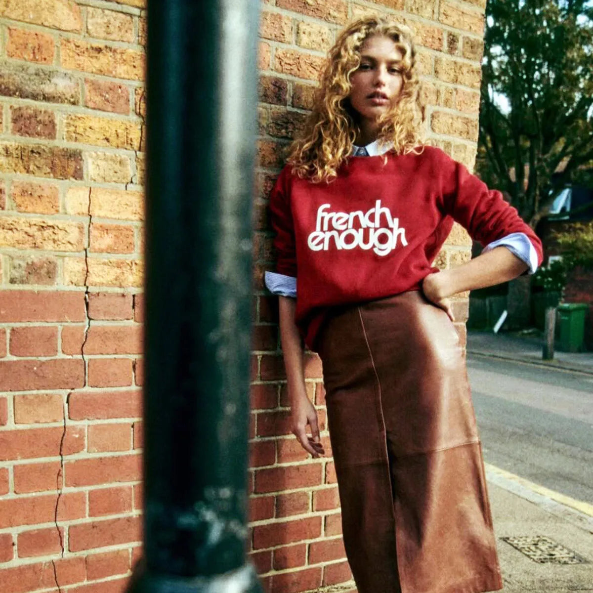 Woman wearing a red sweater with 'French Enough' text against a brick wall.