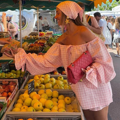 Woman in a pink checkered outfit at an outdoor market with fruit stands.