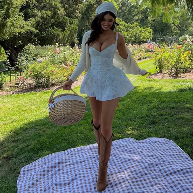 Woman in a light blue dress and white cardigan standing on a checkered blanket in a garden.