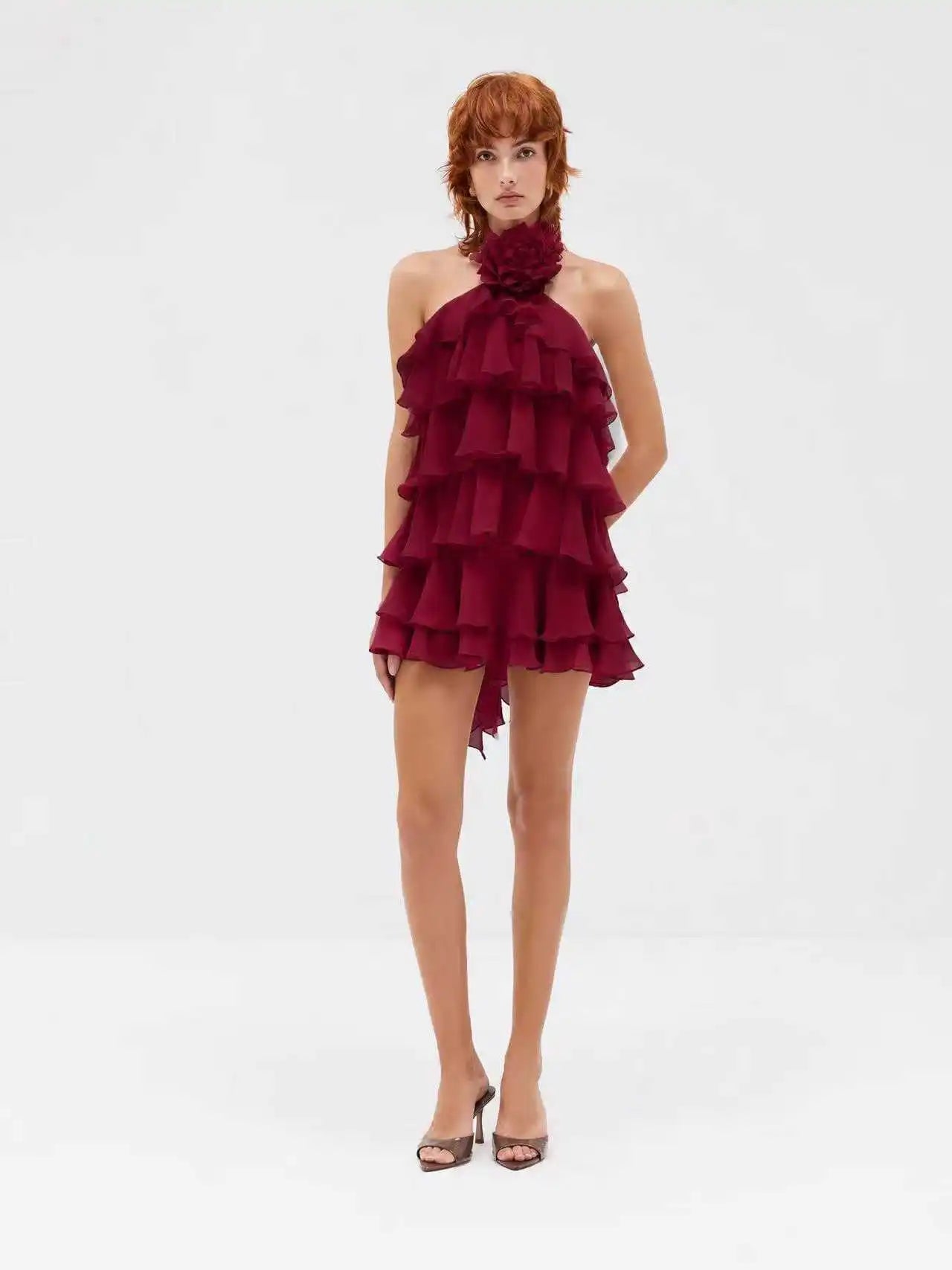 A model wearing a red halter-neck mini dress with dramatic tiered ruffles and a matching fabric flower collar, paired with brown strappy heels, against a plain white studio background.