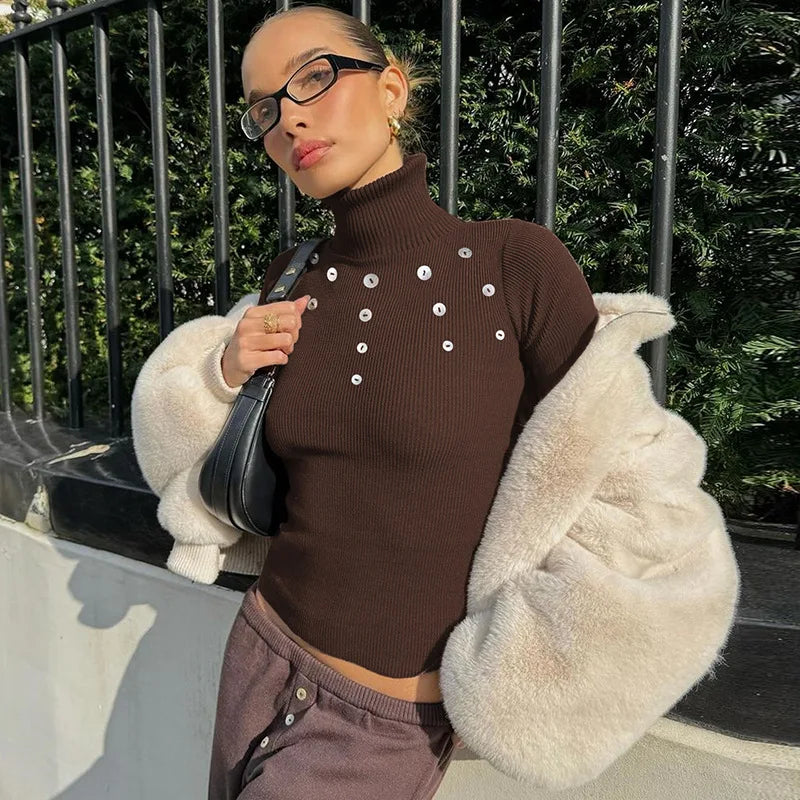 Person wearing a brown turtleneck sweater with silver buttons, holding a black handbag, and wearing a white fur coat.