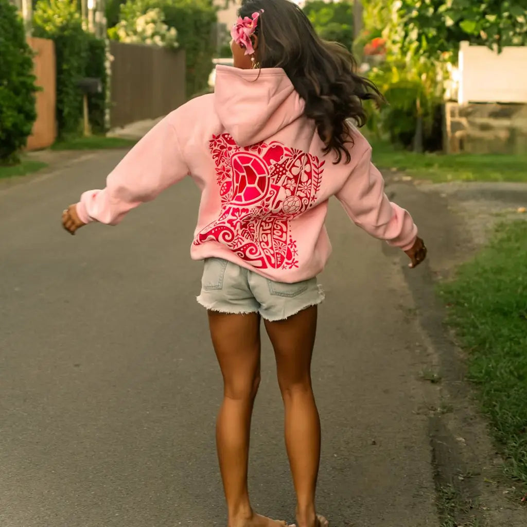 Person wearing a pink hoodie with a red design on the back, standing on a street.