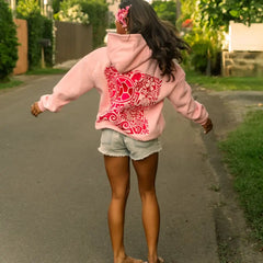 Person wearing a pink hoodie with a red design on the back, standing on a street.