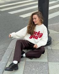 Person sitting on a street corner wearing a white sweater with red text and brown pants.