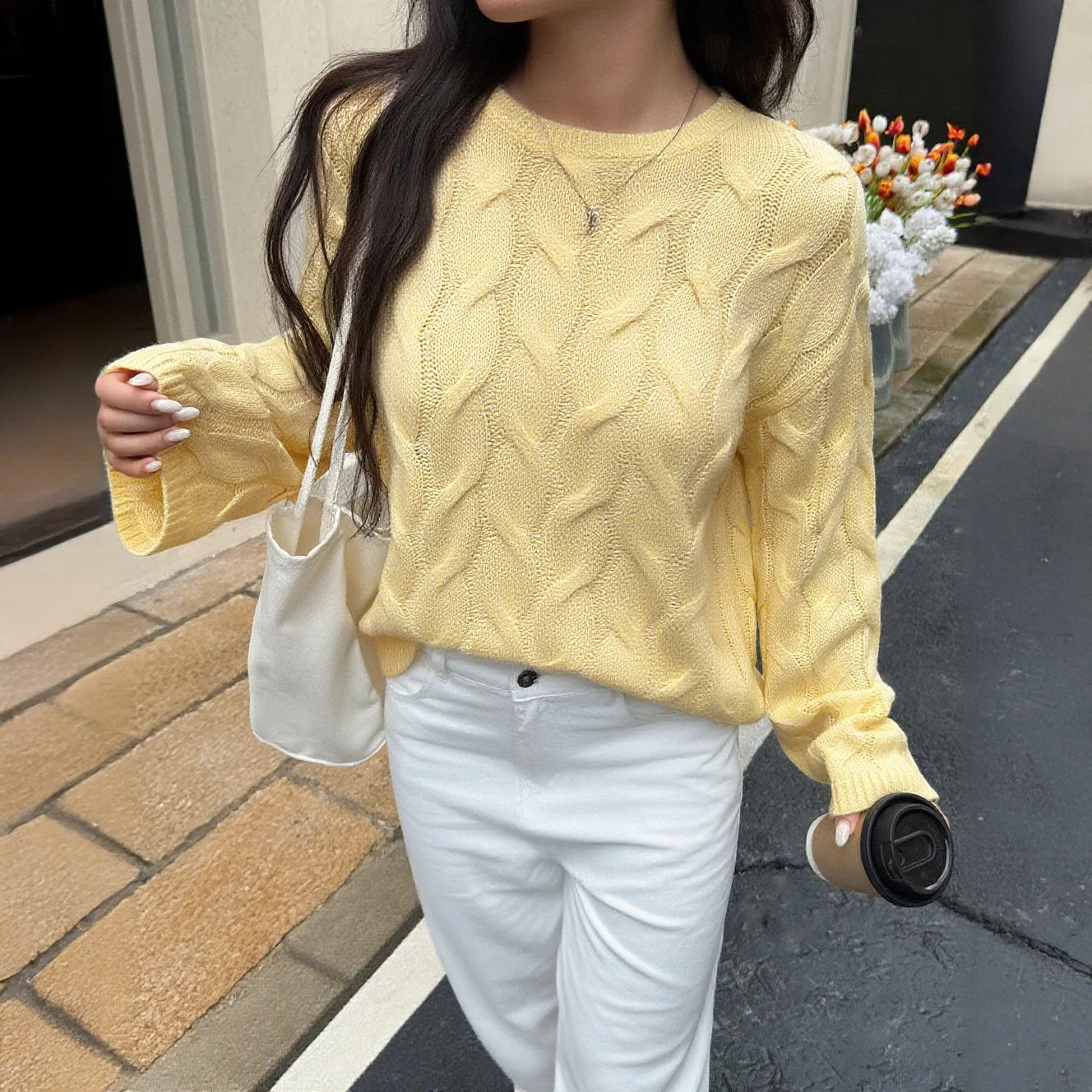 Person wearing a yellow cable knit sweater and white pants, holding a beige handbag and a black coffee cup.