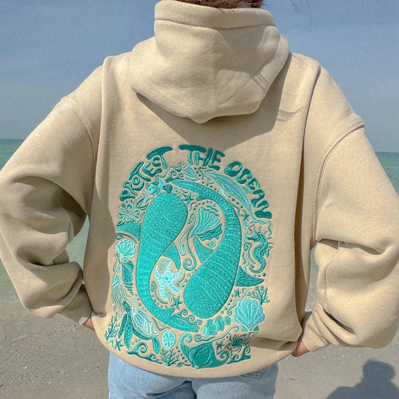 Person wearing a beige hoodie with a teal ocean-themed design on a beach.