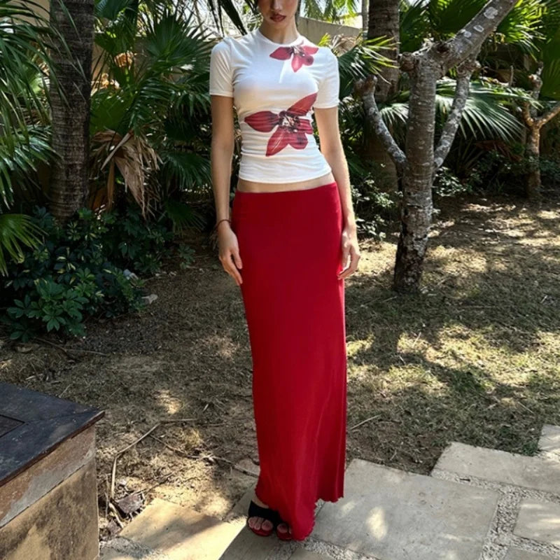 Person wearing a white t-shirt with a floral design and a long red skirt standing in a garden.