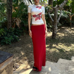 Person wearing a white t-shirt with a floral design and a long red skirt standing in a garden.
