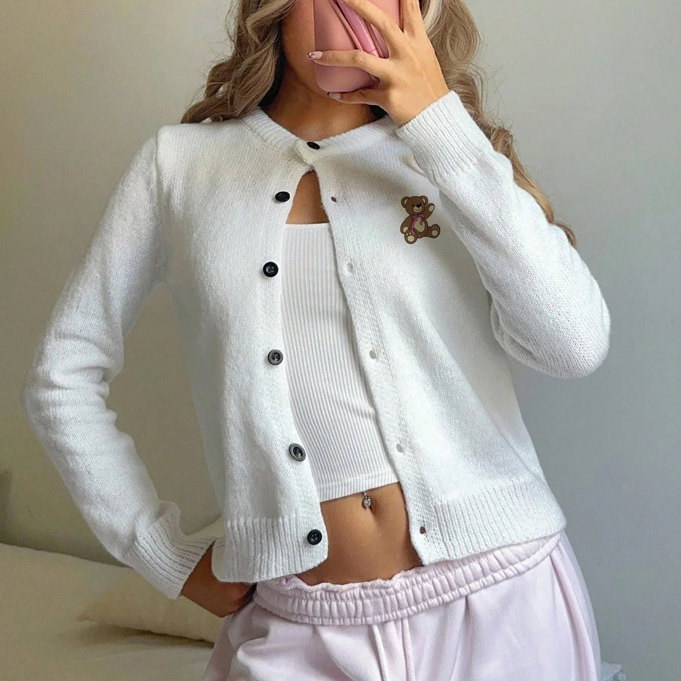 White cardigan with black buttons and a teddy bear emblem worn by a person, sitting on a light-colored surface.