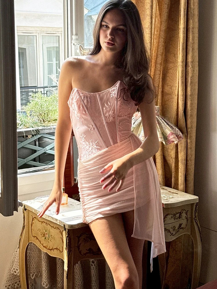 Woman in a pink dress standing indoors with curtains and a window in the background