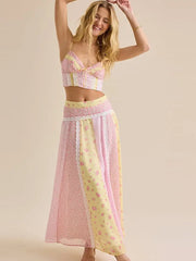 A woman modeling a pastel two-piece outfit consisting of a cropped camisole top and a long maxi skirt, featuring pink and pale yellow floral and lace patterns against a solid beige background.