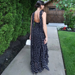 Woman in a black polka dot dress walking on a sidewalk with greenery and a wooden shed in the background.