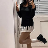 Person wearing a black sweater with a pleated skirt and high-heeled shoes, standing against a white wall.