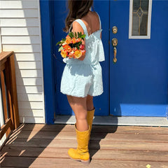 Person in a white dress with yellow boots holding flowers in front of a blue door.