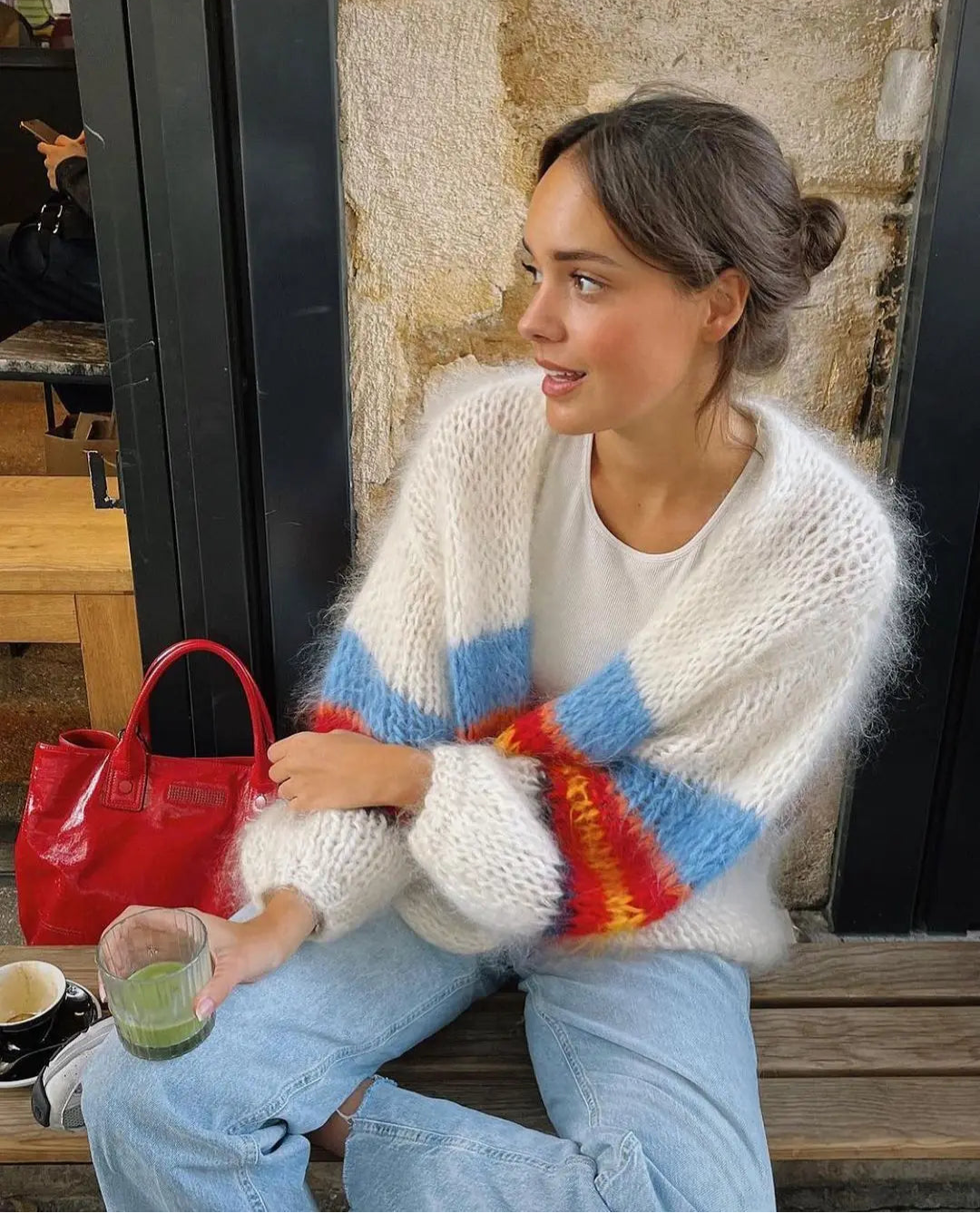 Woman sitting outdoors wearing a colorful striped sweater and holding a red handbag.