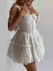 A woman wearing a white eyelet lace mini dress with a corset-style bodice, tiered ruffle skirt, and thin shoulder ties, photographed against a plain light background.
