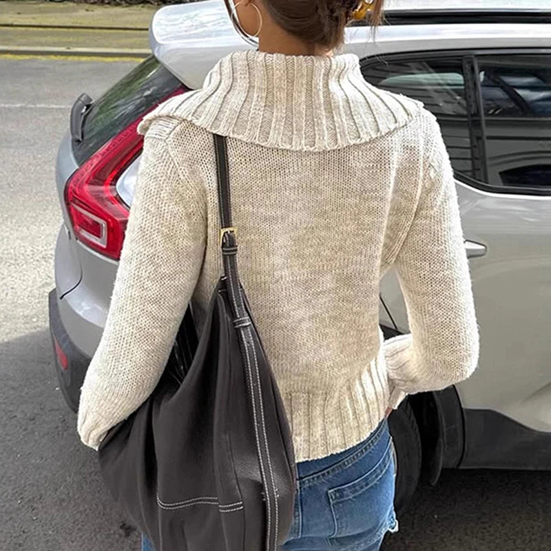 Person wearing a beige sweater and carrying a black bag, standing next to a white car.