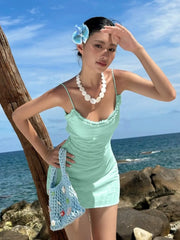 Woman in a light green swimsuit with a blue floral headpiece, standing by the ocean.