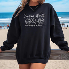 Person wearing a black sweatshirt with 'Cousins Beach' design on a beach.