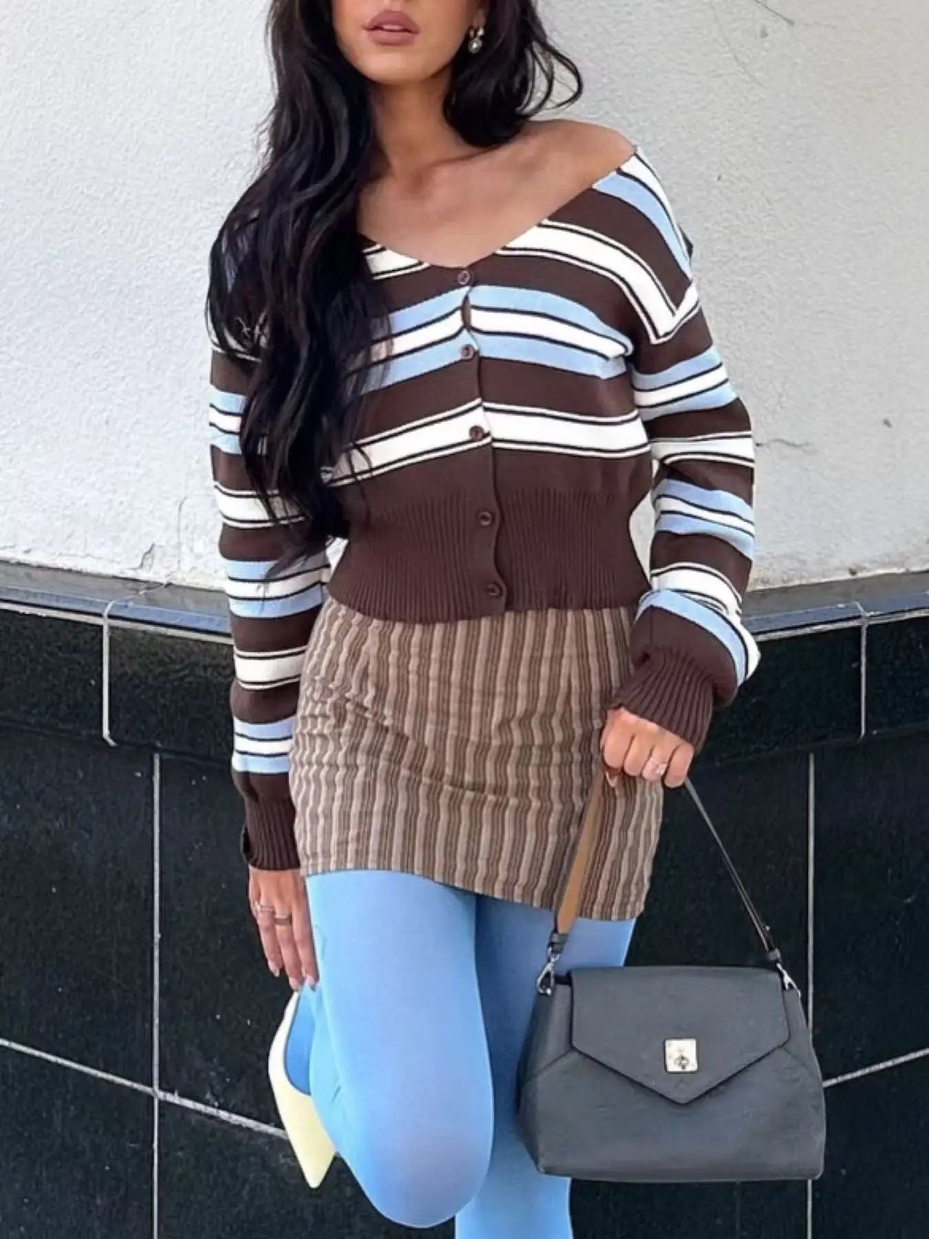 Woman wearing a striped sweater and skirt with a gray handbag against a white wall.