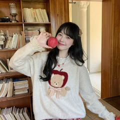 Person wearing a sweater with a teddy bear design, holding an apple indoors.