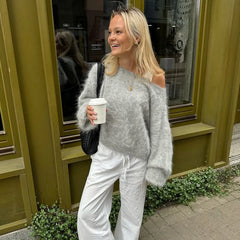 Woman in a gray sweater and white pants holding a coffee cup, standing in front of a green building.