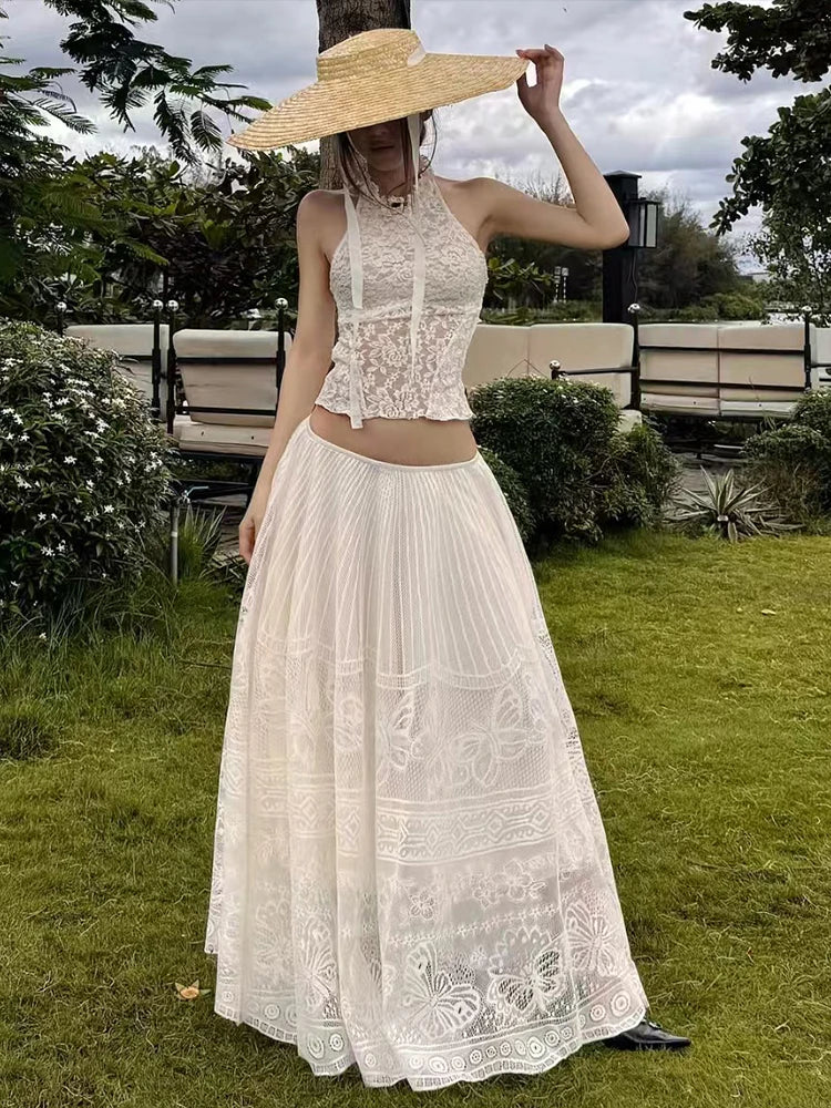 Woman in a white lace dress and wide-brimmed hat standing outdoors on grass with trees and benches in the background.