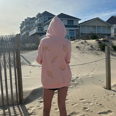Person wearing a pink hoodie with shell designs on a sandy beach with houses in the background.