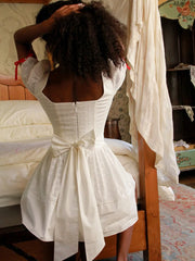 Woman in a white dress standing in a bedroom with wooden furniture and floral patterns- back view