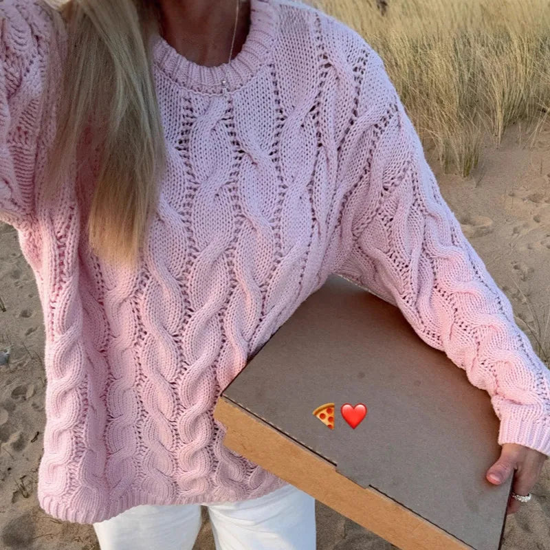 Person wearing a pink knitted sweater holding a pizza box on a beach.