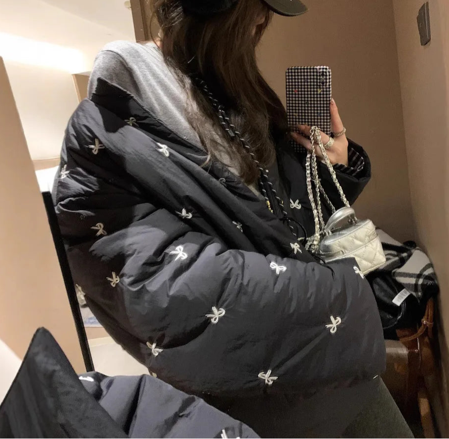 Person wearing a black puffer jacket with white patterns, holding a phone and a handbag indoors.