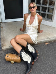 Woman in a white dress and black cowboy boots sitting on a sidewalk.
