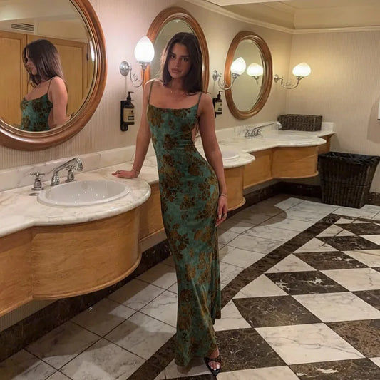 Woman wearing a long, green floral print maxi dress with spaghetti straps and an open back, leaning against a marble countertop in a bathroom.