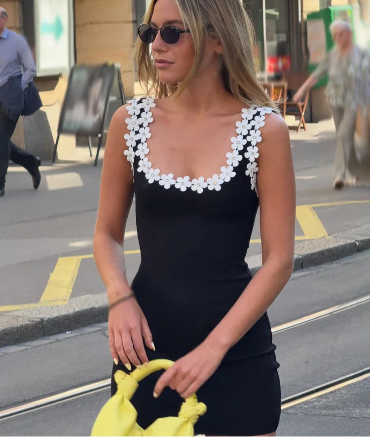 A woman wearing a black mini dress with white floral appliques on the shoulder straps, holding a yellow bag.