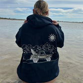 Person wearing a navy hoodie with a graphic design of a beach scene, standing by a body of water.