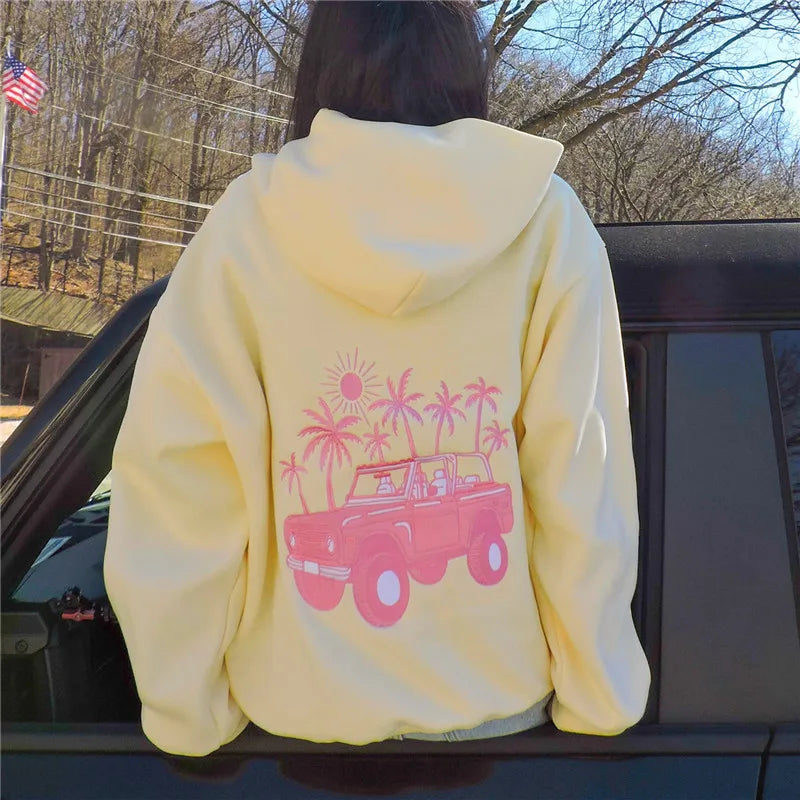 Person wearing a yellow hoodie with pink graphic design, standing next to a car outdoors.
