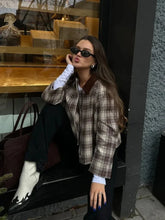 Woman in plaid jacket and white boots leaning against a wall with a store window in the background