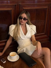 Woman in a white outfit sitting at a table with a cup of coffee and a phone, against a wooden panel background.