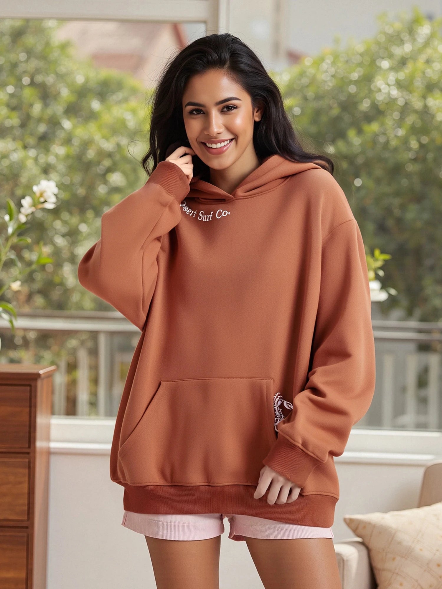 Woman wearing a brown hoodie with visible branding, standing indoors with a blurred background
