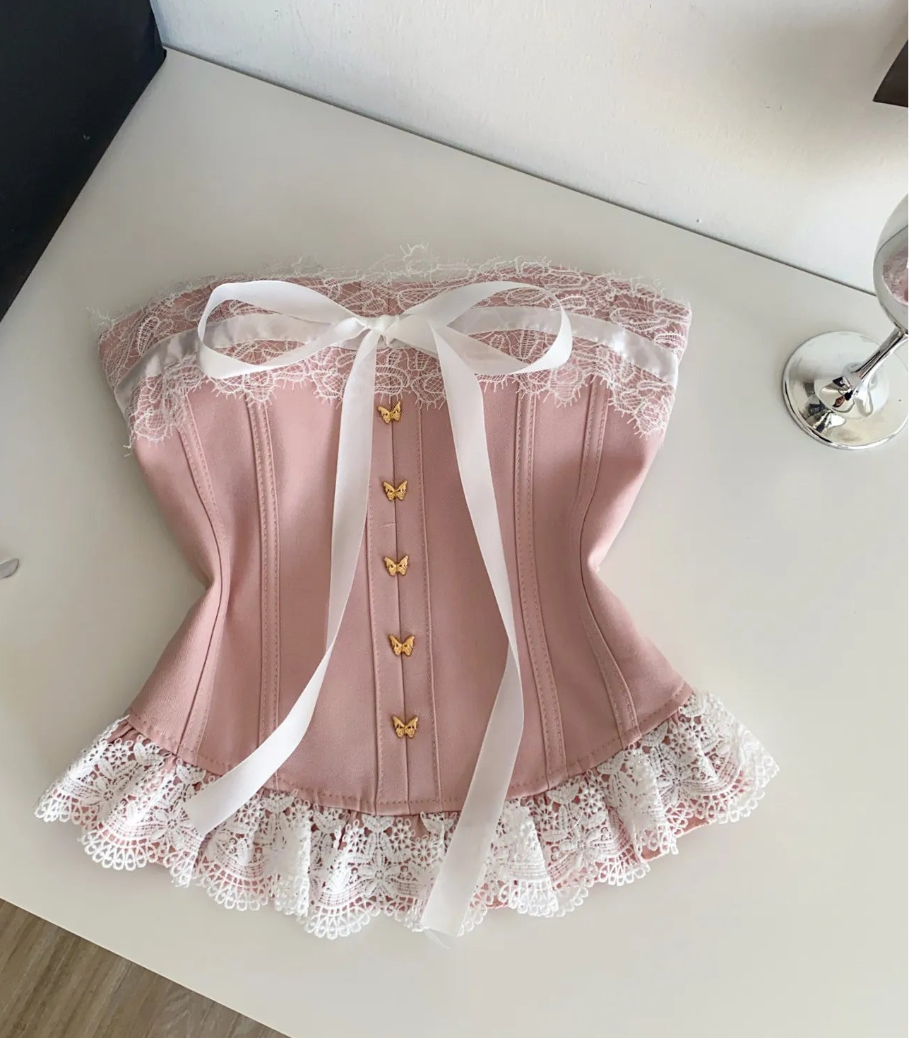 A pink lace-trimmed corset top with a white ribbon bow and gold butterfly details, laid flat on a white tabletop with a Chanel box, sunglasses, and a wine glass nearby.