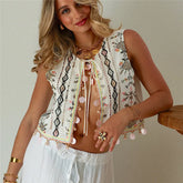 Woman wearing a patterned top with pom-pom details and white pants against a neutral background