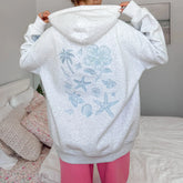 Person wearing a white hoodie with blue ocean-themed designs, standing in a bedroom.