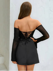 Black off-shoulder dress with lace-up back worn by a person against a white wall.