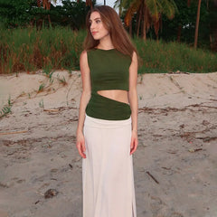Woman wearing a green top and white pants standing on a sandy beach with grass and palm trees in the background.